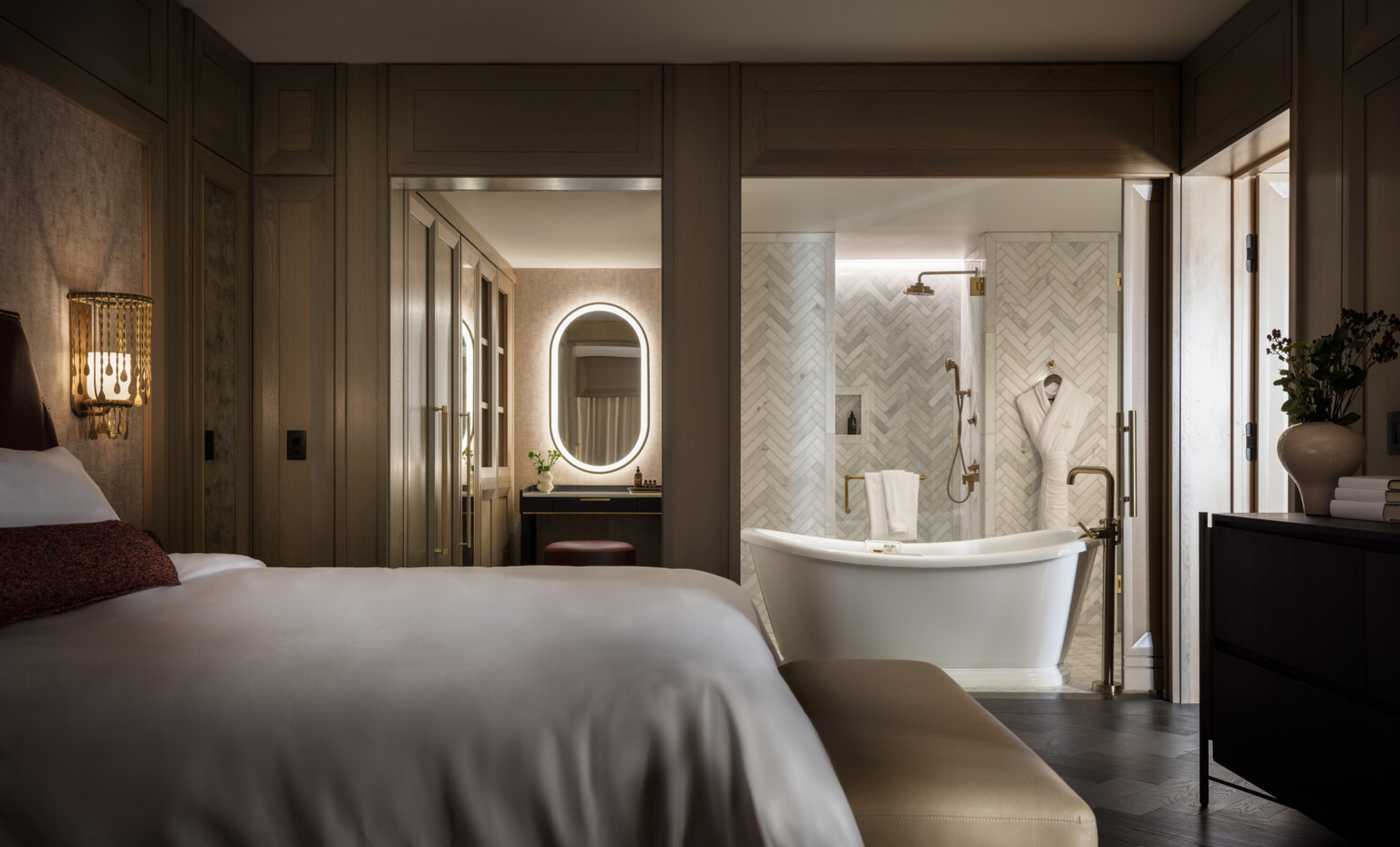 Dimly lit hotel room showcasing a bathroom with large tub in front of a grey and white tile wall, oval lit mirror, bed with soft white linens