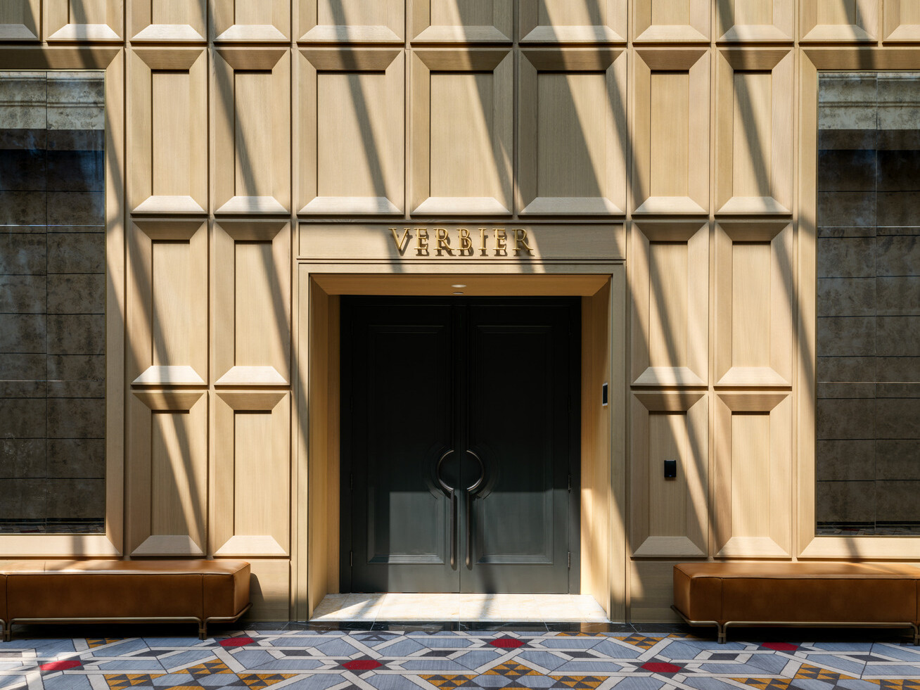 Doorway with black doors surrounded by light wooden wall filled with rectangular shapes, ceiling filled with windows above allowing natural light into the space