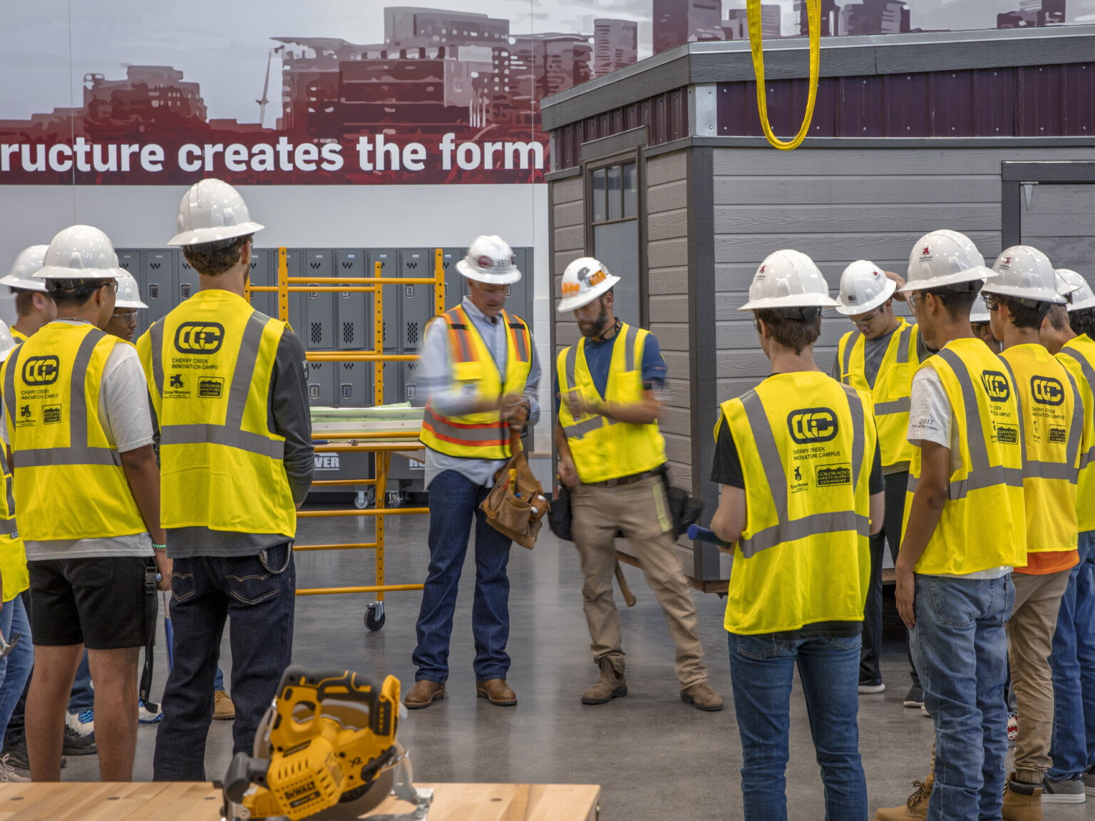 Circular saw on table in front of students in yellow safety vests and hard hats circled around teachers. Paneled building in back right