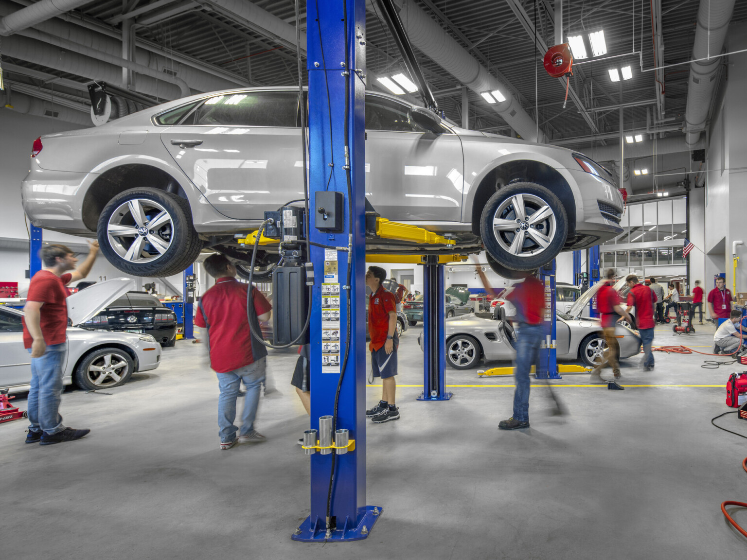 Students work on lifted car in double height mechanics lab. More cars on the ground behind. Exposed duct work on grey ceiling