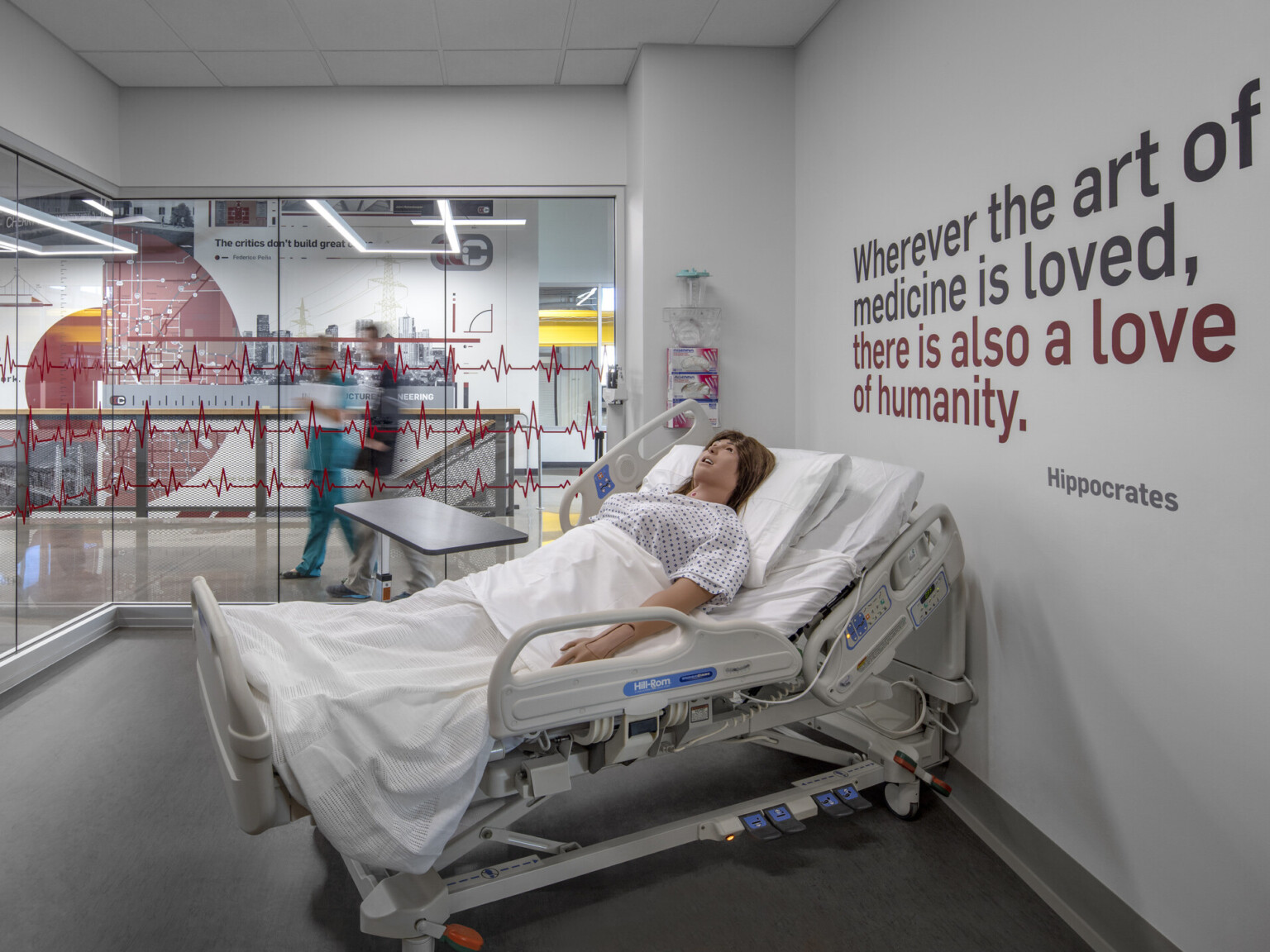 Health science lab with hospital bed and CPR dummy. Hippocrates quote on right wall. Glass walls to hall with zigzag design