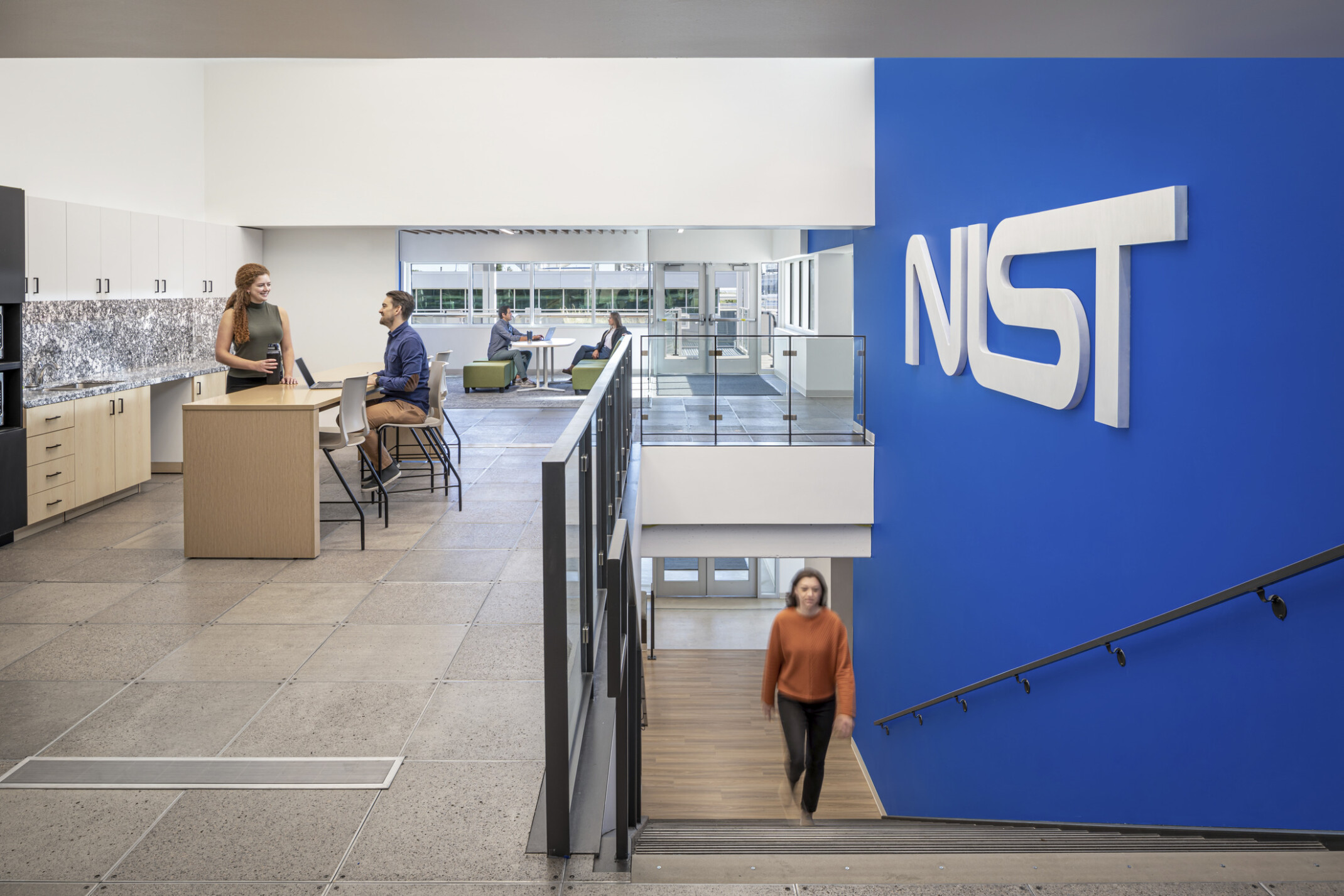 Woman in a orange shirt walking up a staircase with a blue wall to the right with letters NIST. Two couples at the top of the stairs working in collaborative areas; NIST Boulder