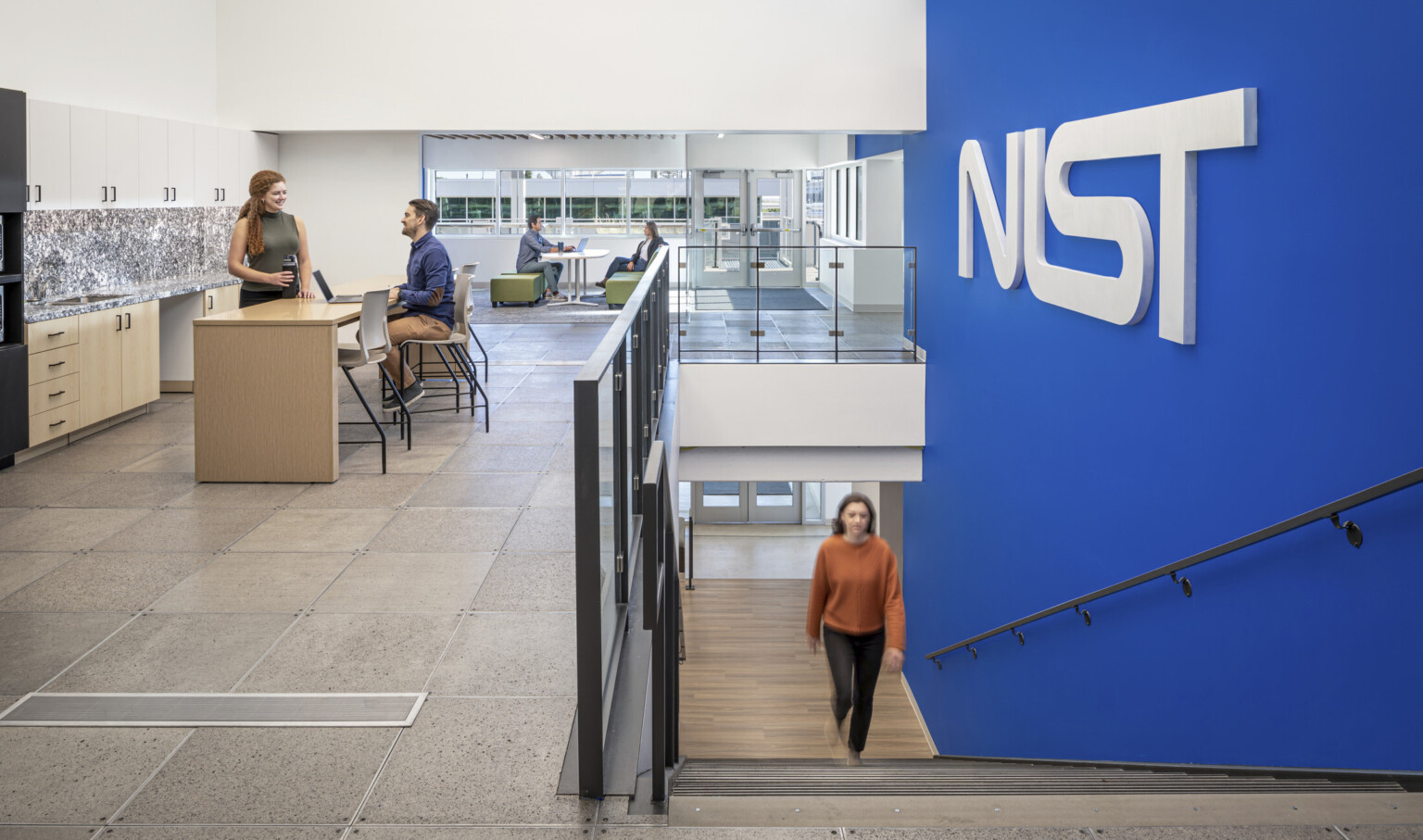 Woman in a orange shirt walking up a staircase with a blue wall to the right with letters NIST. Two couples at the top of the stairs working in collaborative areas; NIST Boulder