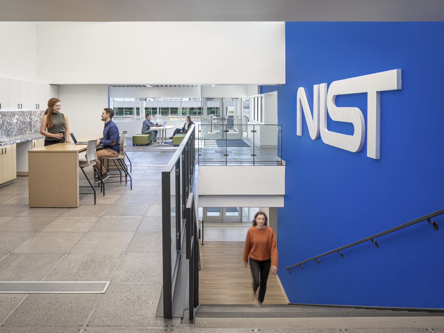 Woman in a orange shirt walking up a staircase with a blue wall to the right with letters NIST. Two couples at the top of the stairs working in collaborative areas; NIST Boulder