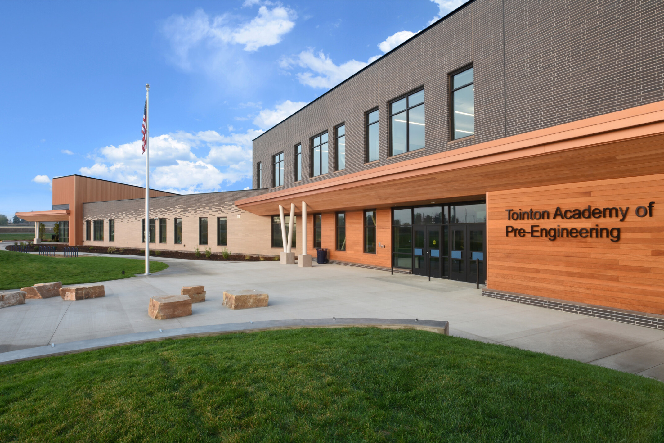 Exterior of two-story engineering academy entrance, walkway, grass, outdoor seating, double-height windows, flag pole with American flag, natural rock seating, under blue skies and fluffy white clouds