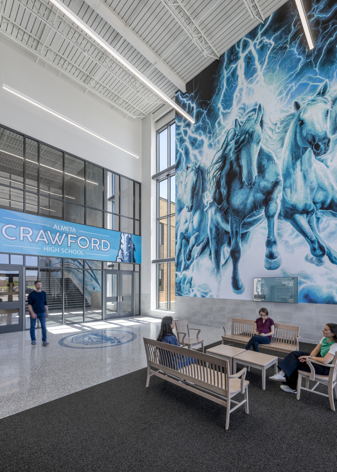 Seating area with large mural in shades of blue behind benches. Large wall filled with windows with artwork with words Almeta Crawford High school in the middle