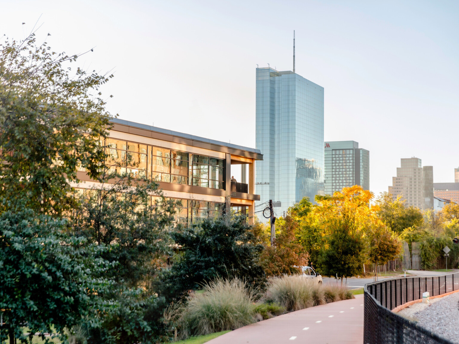 A modern building with glass windows reflects sunlight, surrounded by greenery. A pathway leads to skyscrapers, blending urban life with nature.