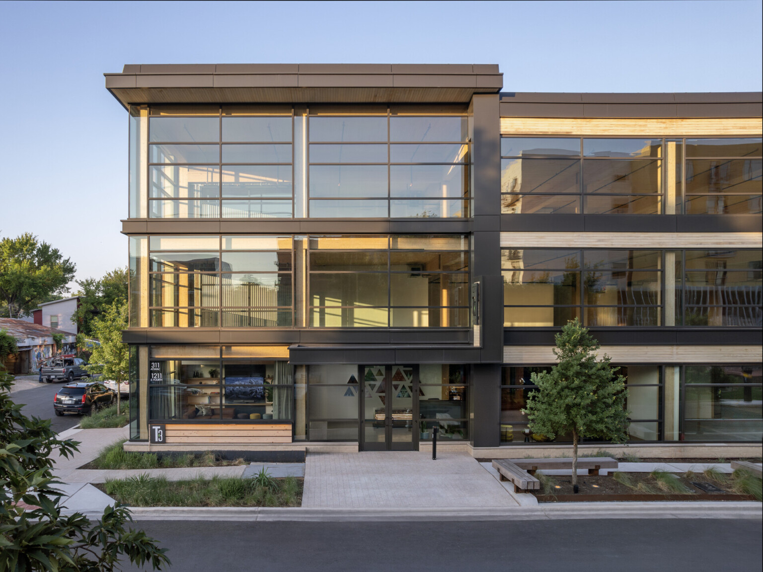 Corner view of building with black exposed steel at front, white ribbed panel, back, exposed mass timber and large windows, greenery