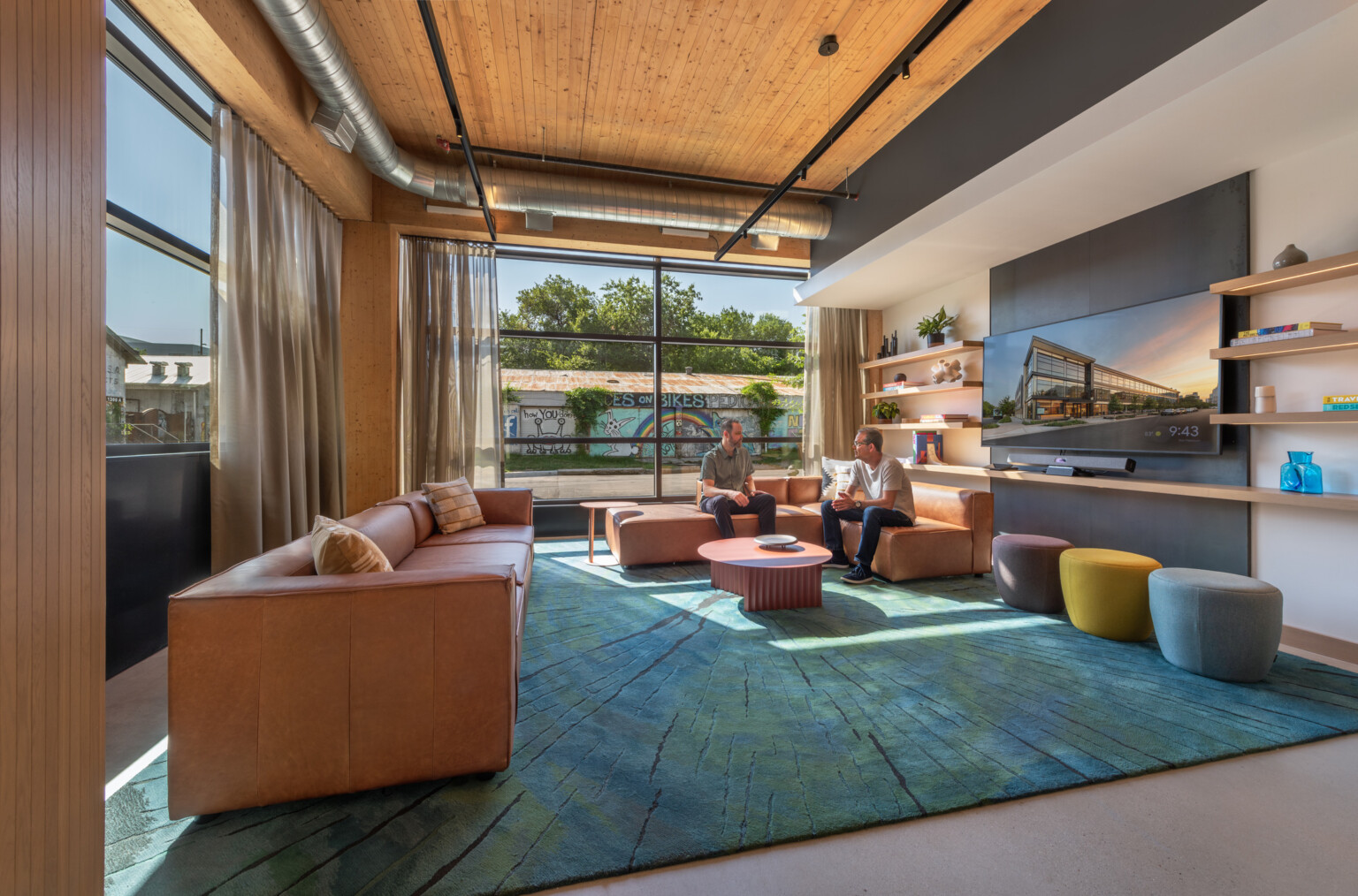 Modern seating area filled with brown leather couches, rug in shades of blue, ottomans, walls with shelves and TV and a colorful mural outside the windows