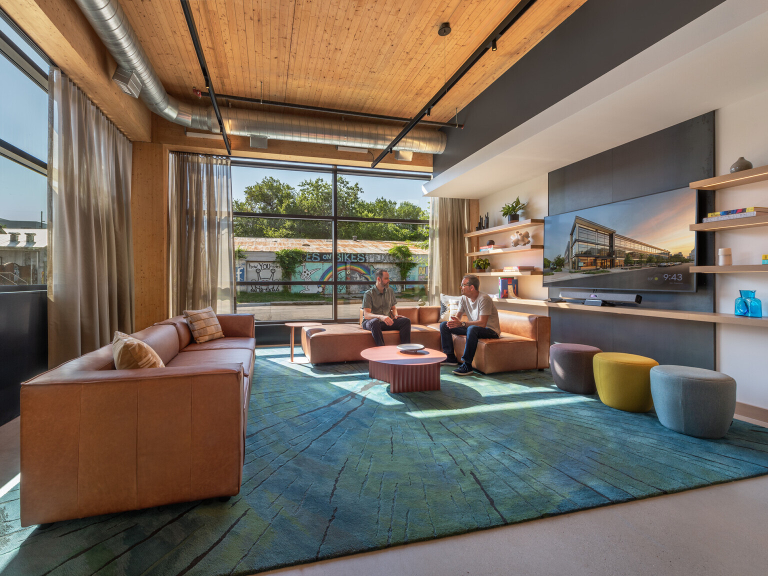 Modern seating area filled with brown leather couches, rug in shades of blue, ottomans, walls with shelves and TV and a colorful mural outside the windows