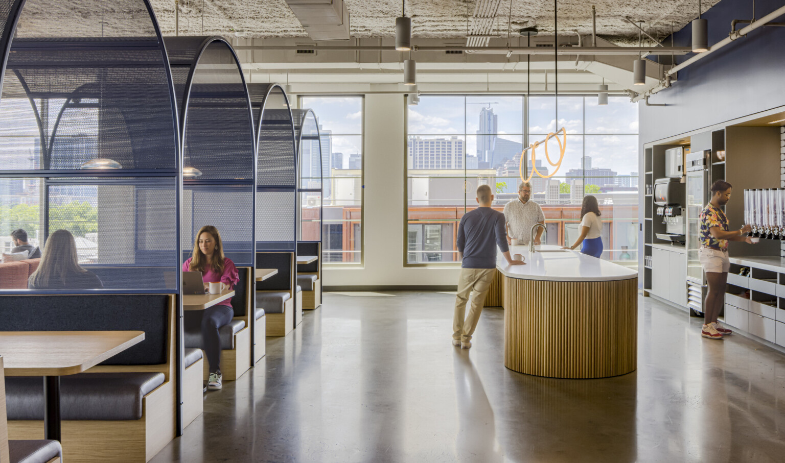 A modern office with booth seating, open spaces, and city views. Employees collaborate near a counter, with an industrial-style ceiling above.