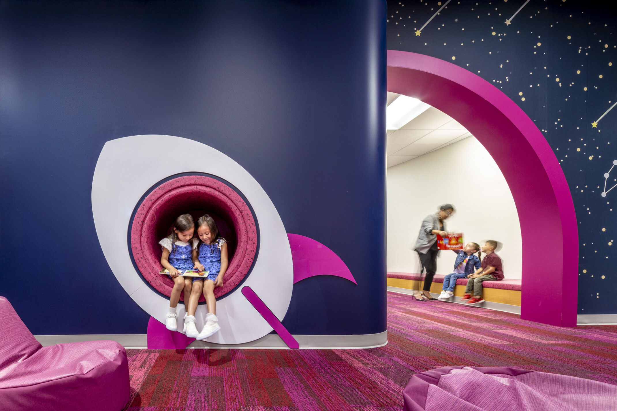 kids reading a book in a cutout window of a space ship mural. Blue walls with pink accents at a early excellence academy.