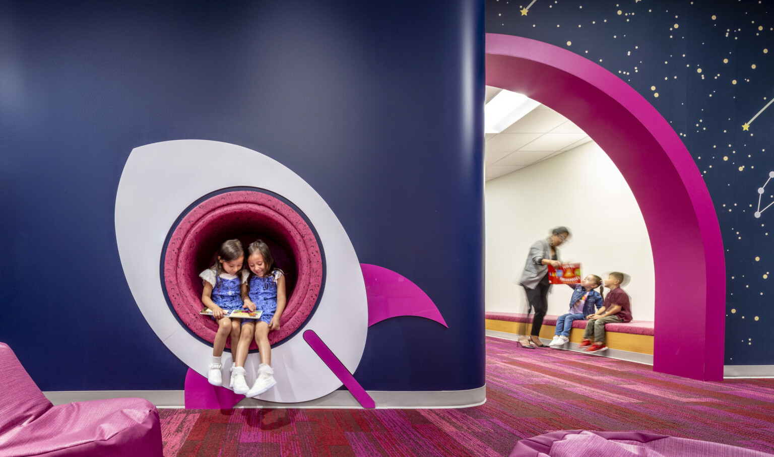 kids reading a book in a cutout window of a space ship mural. Blue walls with pink accents at a early excellence academy.