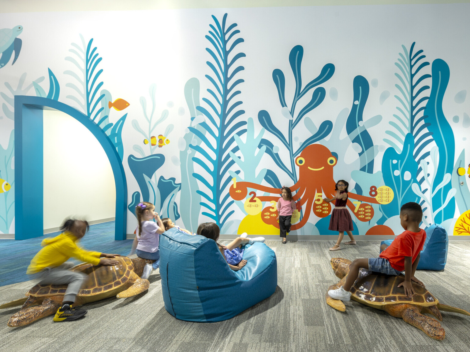 Kids interacting at an early excellence academy in front of a colorful ocean wall mural in shades of blue with an orange octopus surrounded by orange and yellow fish.