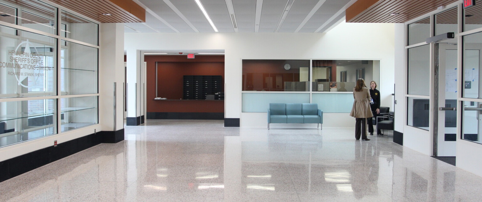 Office hallway surrounded by glass windows looking at a blue couch on a wall with multiple windows