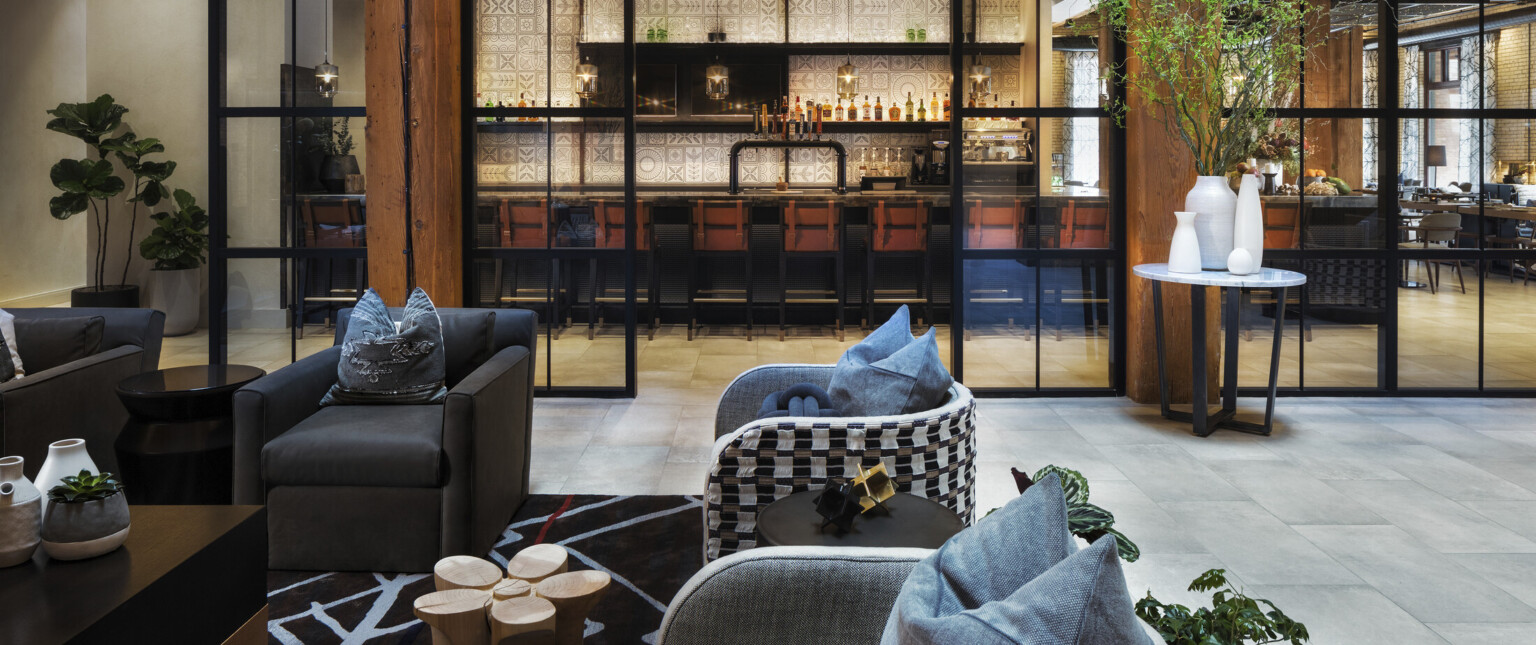 Seating area in room with double height windows looking to white geometric walled bar in next room. Wood columns and ceiling