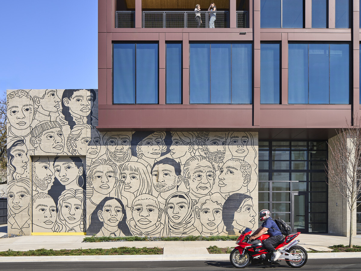 Vertical shot of a multistory mass timber building with rows of windows of floor to ceiling windows. Balconies overlooking the street on multiple floors. Large mural of faces at street level