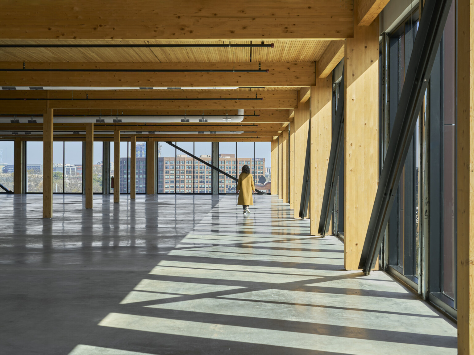 Interior of a mass timber office building showing an open floor with exposed wooden accents on the ceiling and wall beams. Large floor to ceiling windows line the exterior of the space. Woman in a yellow coat walking toward the back windows