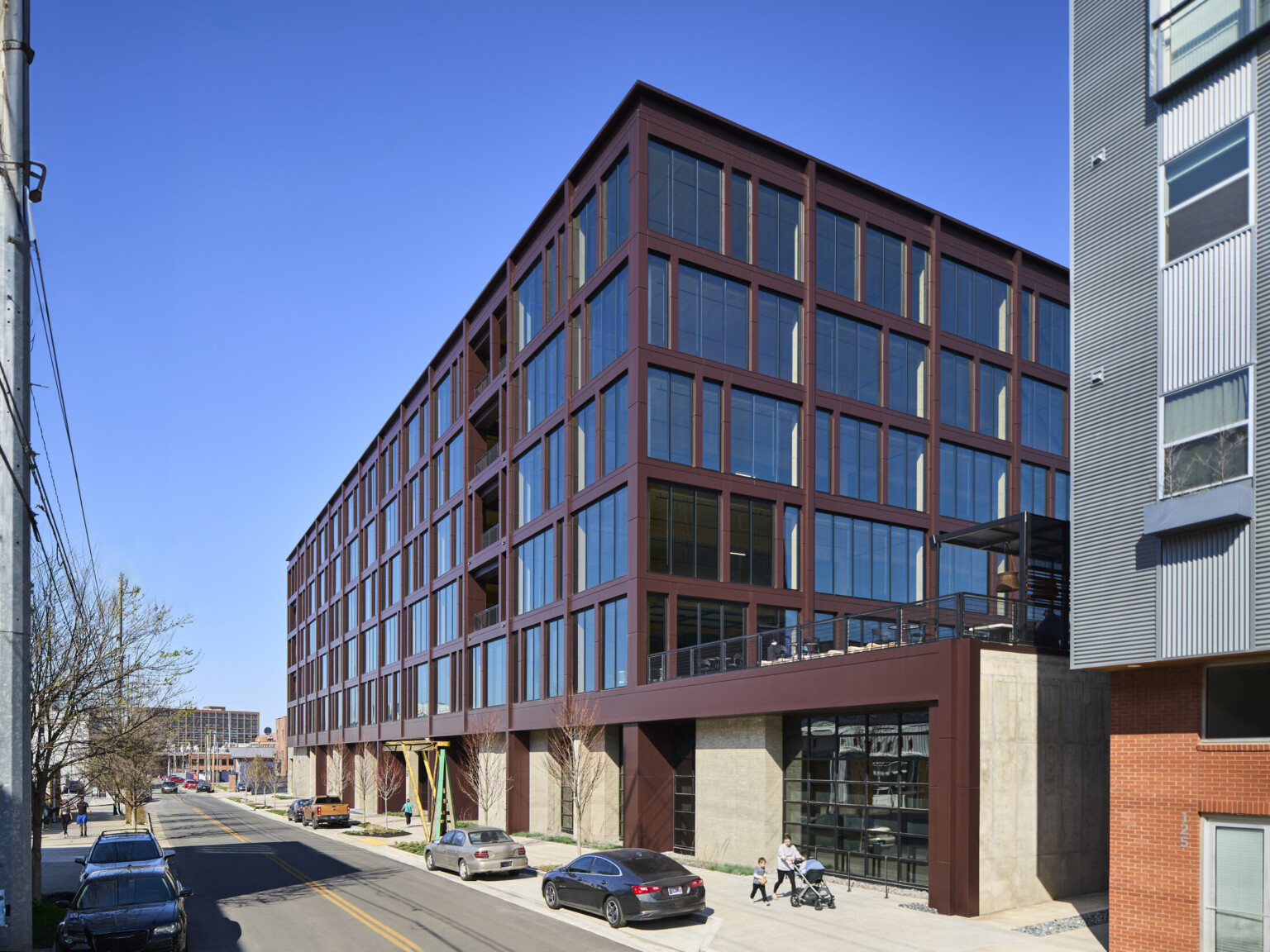 Multistory mass timber building with rows of windows of floor to ceiling windows along a two way road with cars parked on the street