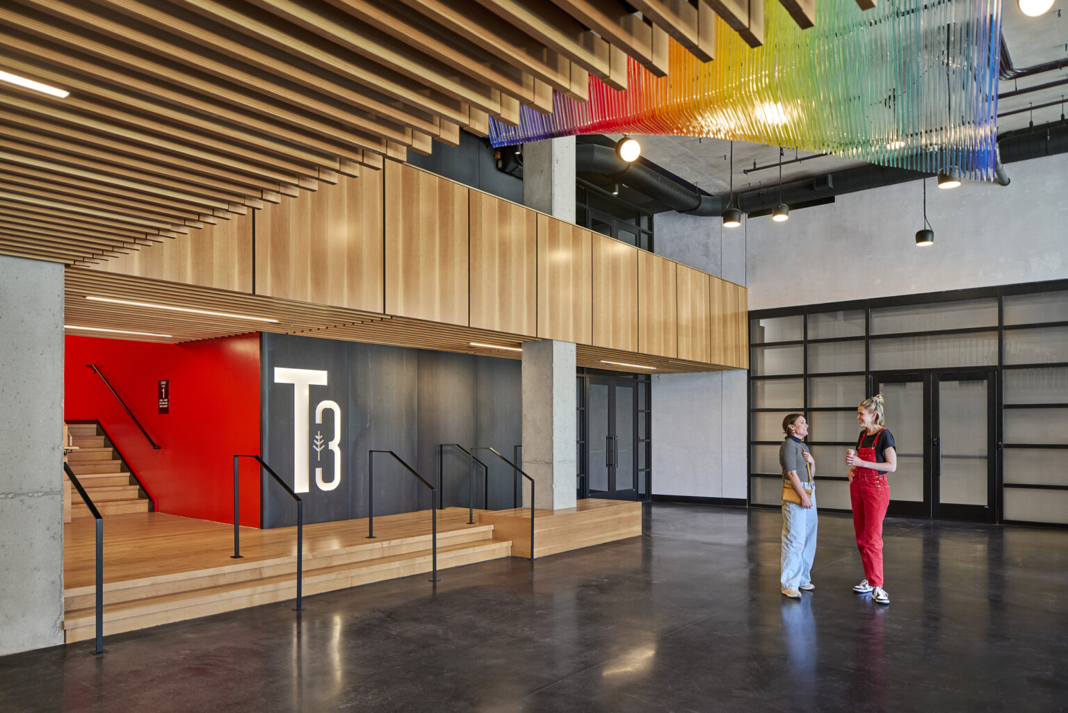 Interior lobby of a mass timber office building showing natural wood accents on the walls, large backlit T3 on back wall and modern rainbow light fixture suspended from the ceiling