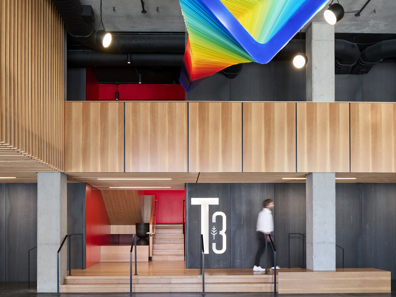 Interior lobby of a mass timber office building showing natural wood accents on the walls, large backlit T3 on back wall and modern rainbow light fixture suspended from the ceiling