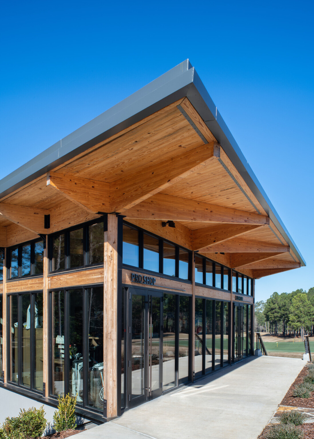 Building made from wood with large floor to ceiling windows outlined in black trim with a large concrete sidewalk in front and large trees in the distance