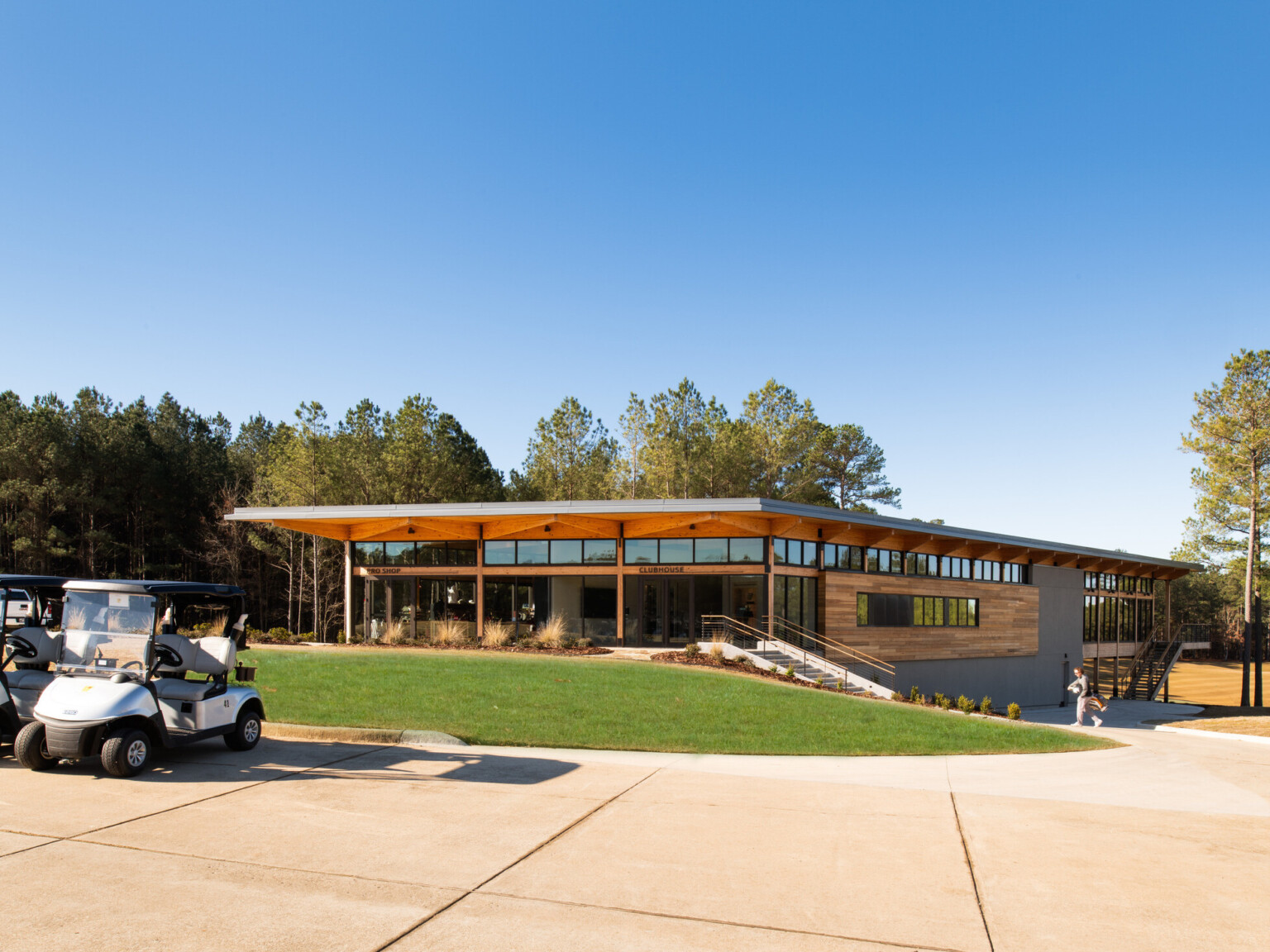 Golf clubhouse and pro shop with wooden accents throughout. Golf carts parked out front with lush grass and large trees in the distance