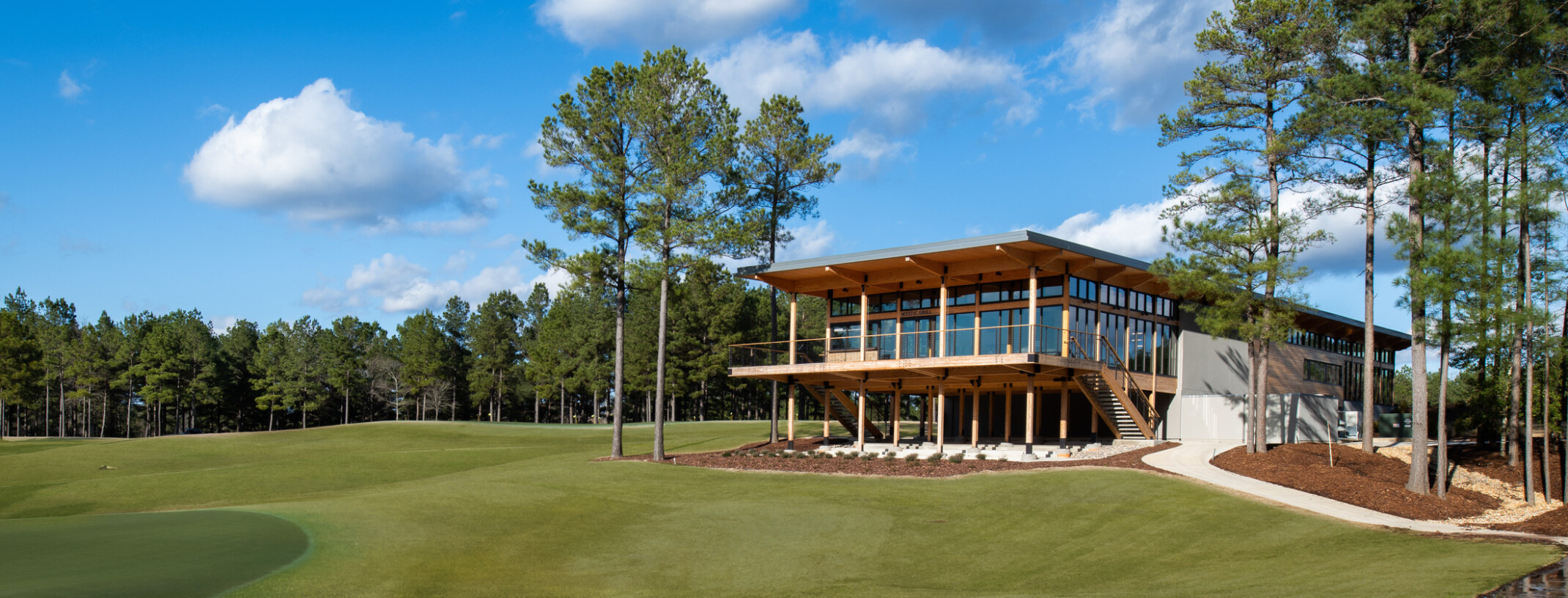 Wooden building supported by pillars on bottom floor. Second floor has floor to ceiling windows with large deck overlooking lush grass and surrounded by trees