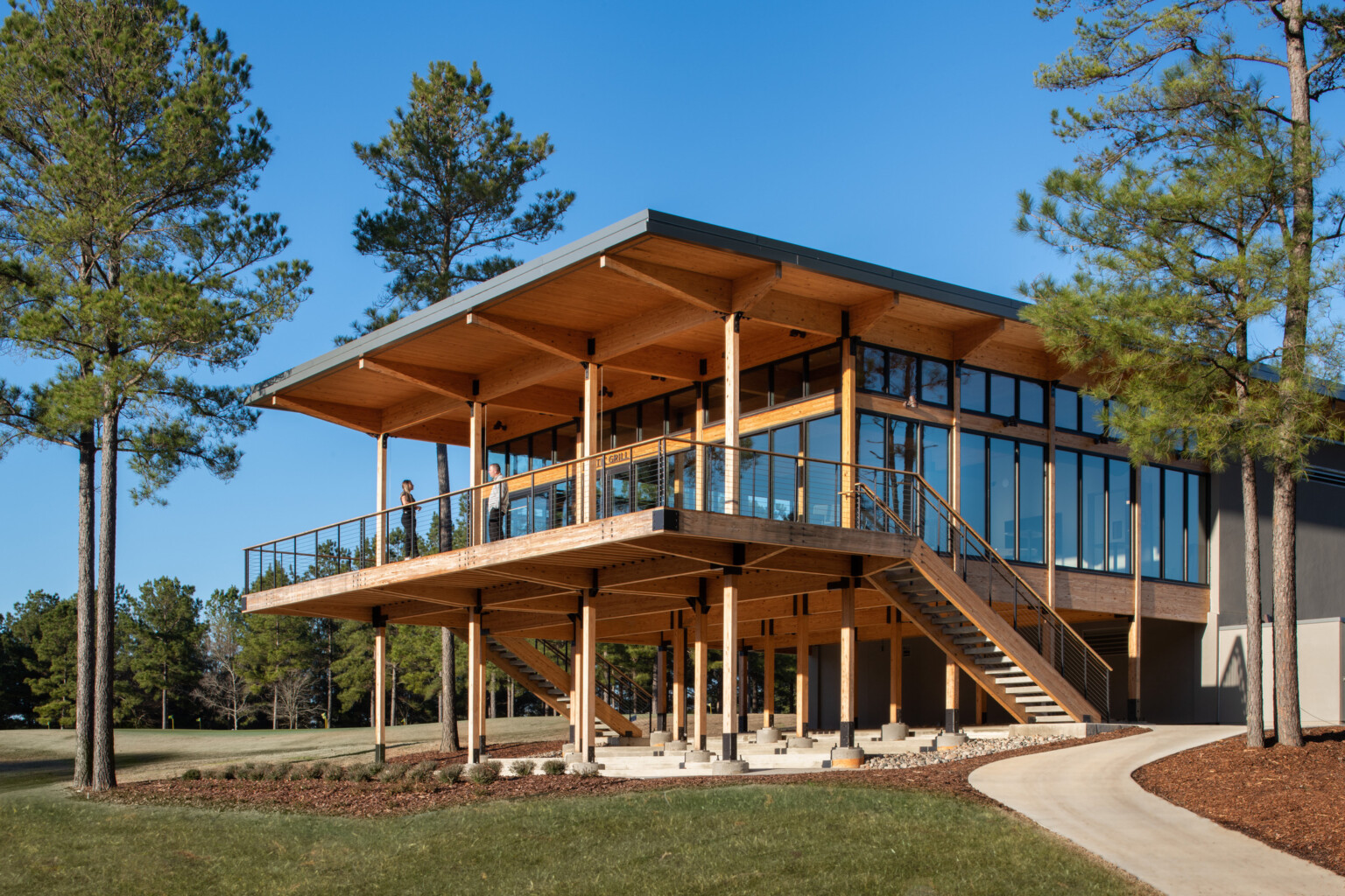 Wooden building supported by pillars on bottom floor. Second floor has floor to ceiling windows with large deck overlook lush grass and surrounded by trees