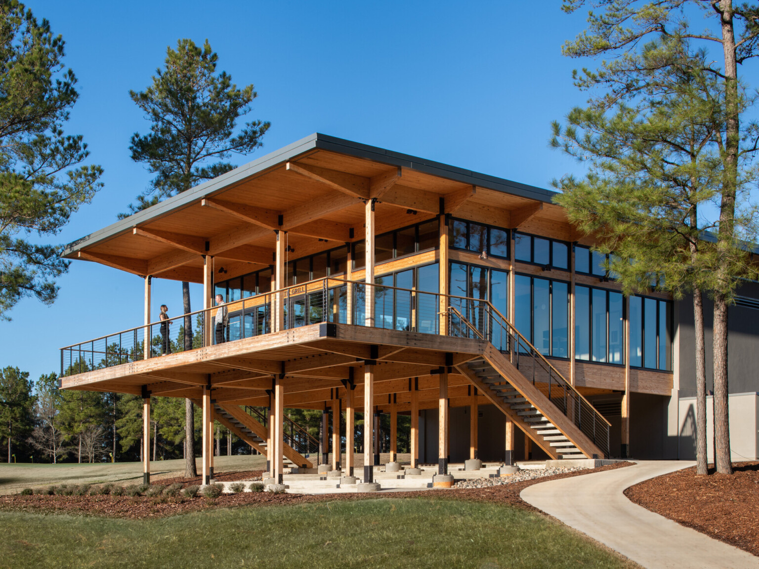 Wooden building supported by pillars on bottom floor. Second floor has floor to ceiling windows with large deck overlook lush grass and surrounded by trees