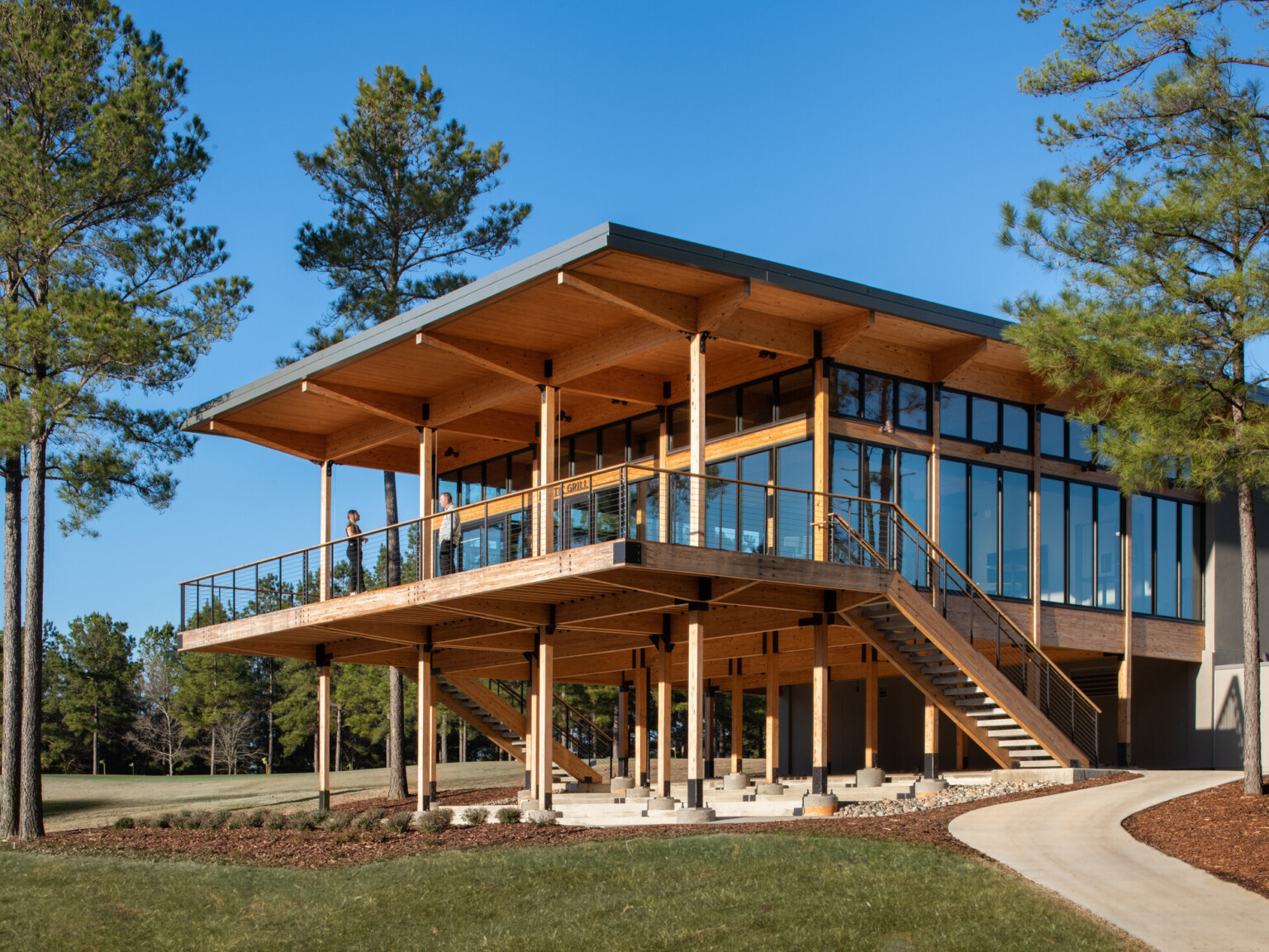 Wooden building supported by pillars on bottom floor. Second floor has floor to ceiling windows with large deck overlook lush grass and surrounded by trees