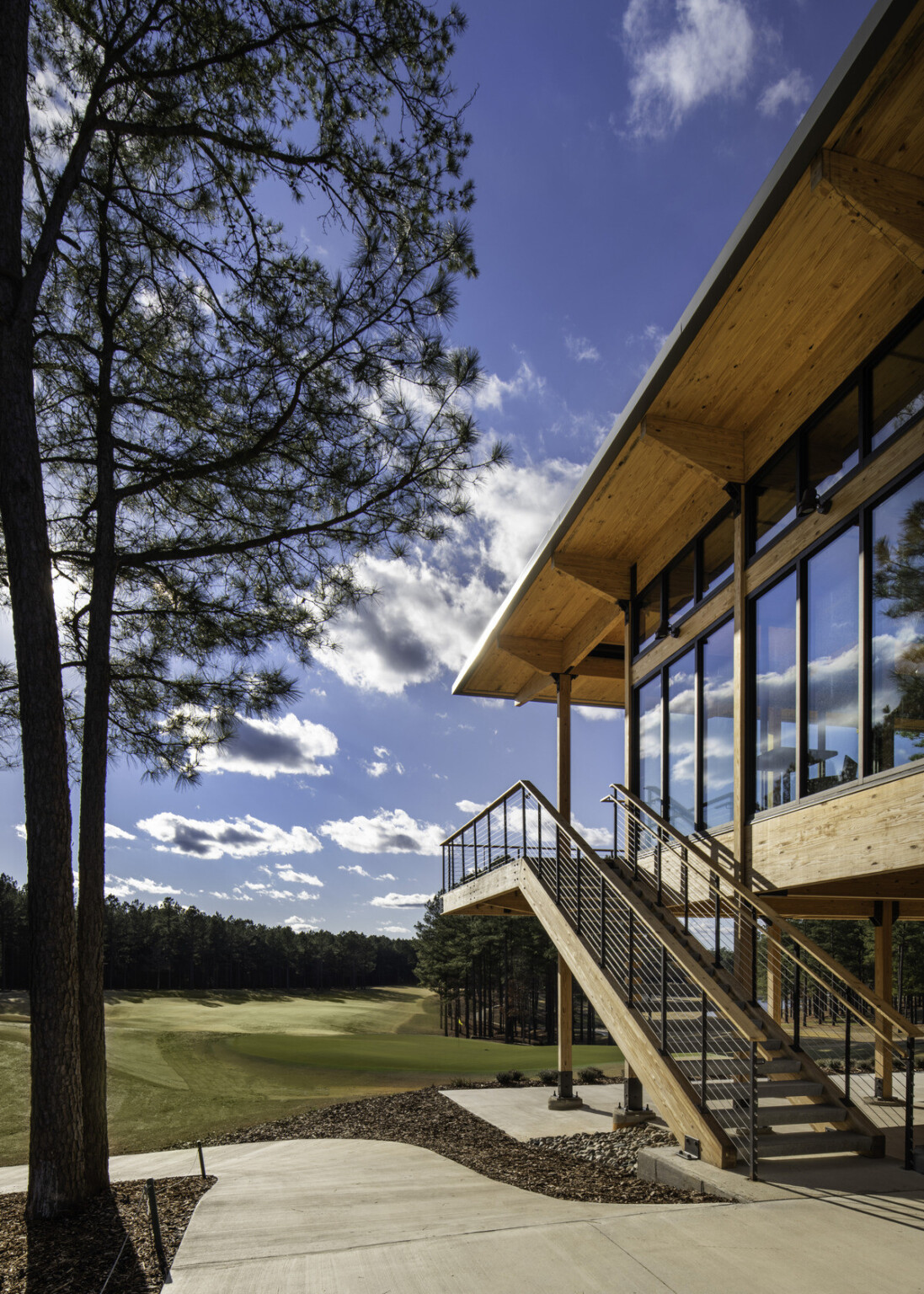 Looking down a golf fairway surrounded by pine trees with a building to the right made from exposed wood with a large wooden staircase with black railings