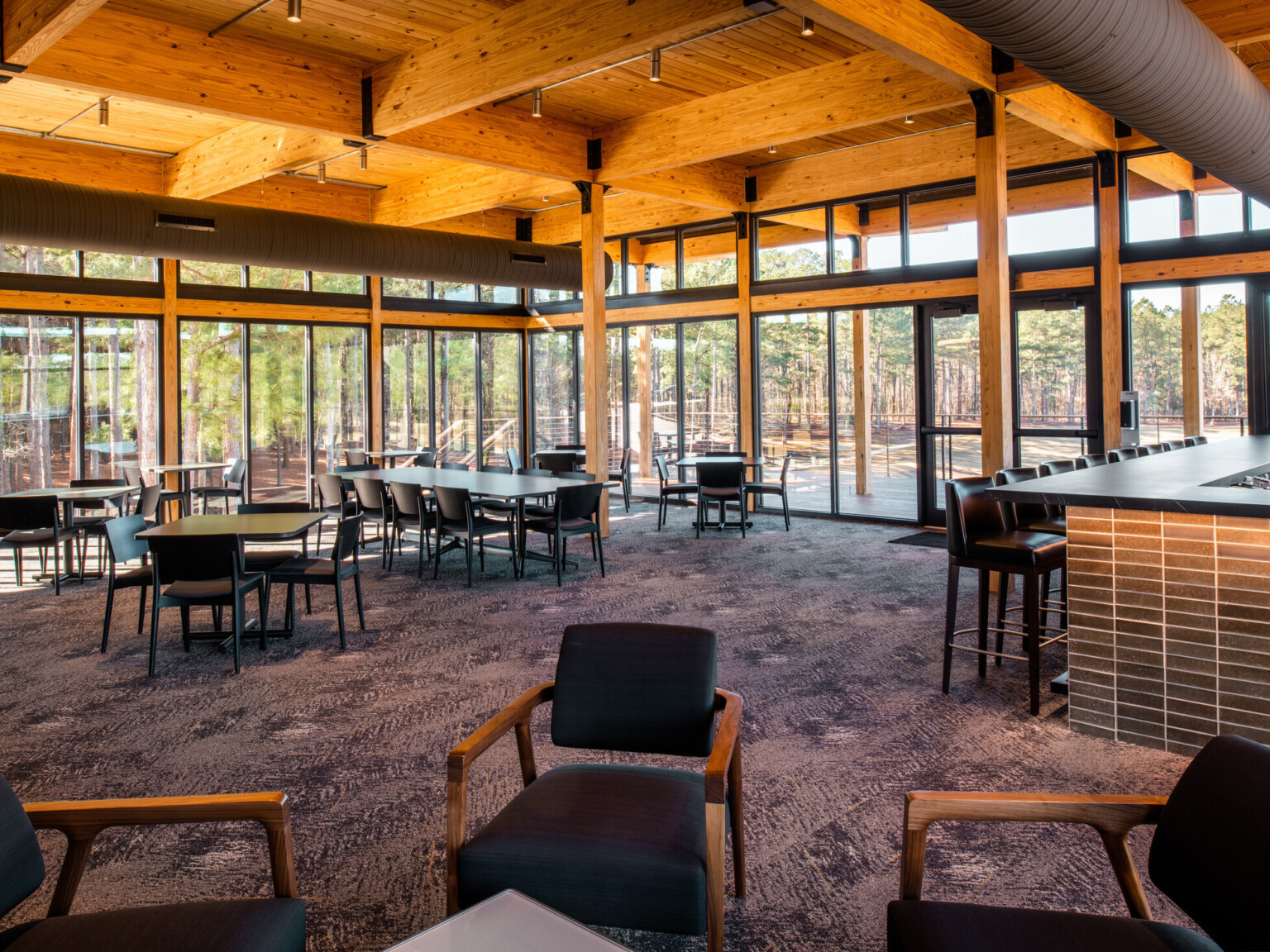 Large dining room filled with black tables and chairs. Exposed wood beams and ceiling throughout with large floor to ceiling windows overlooking large trees in the distance.