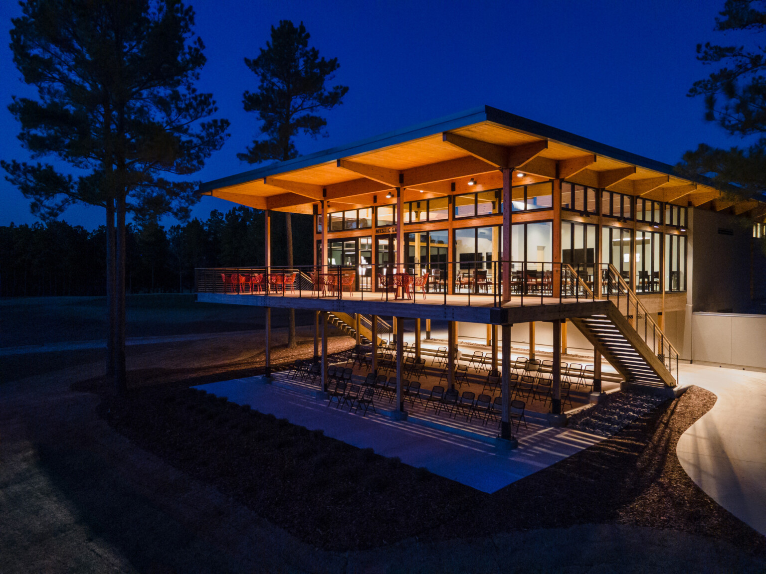 Nighttime shot of a large wooden building supported by pillars on bottom floor. Second floor has floor to ceiling windows filled with light with large deck and trees in the distance