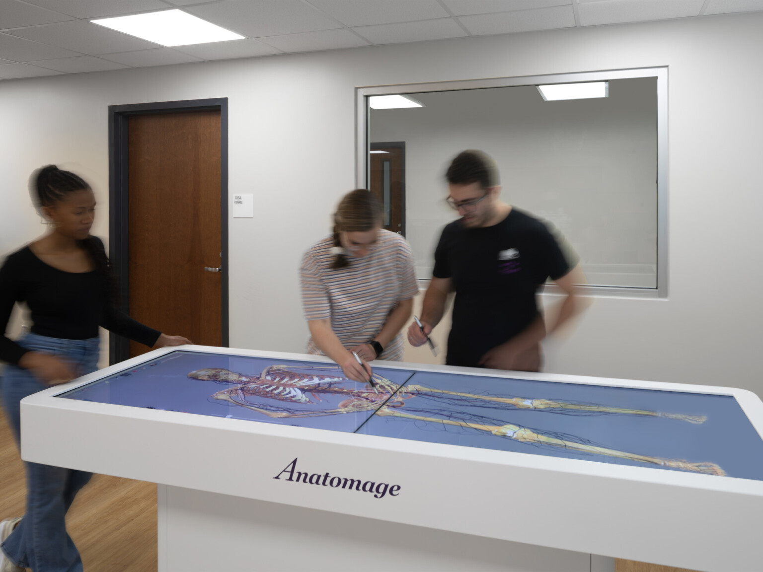 Students surrounding a table with a blue background with a skeleton image on the table with words Anatomage on the side