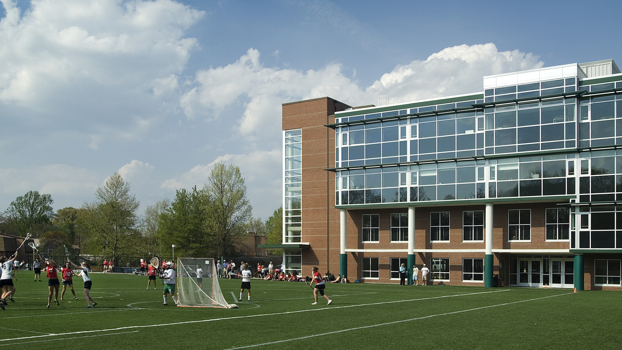 Multistory brick building with glass windows behind a lush green lacrosse field filled with students playing lacrosse