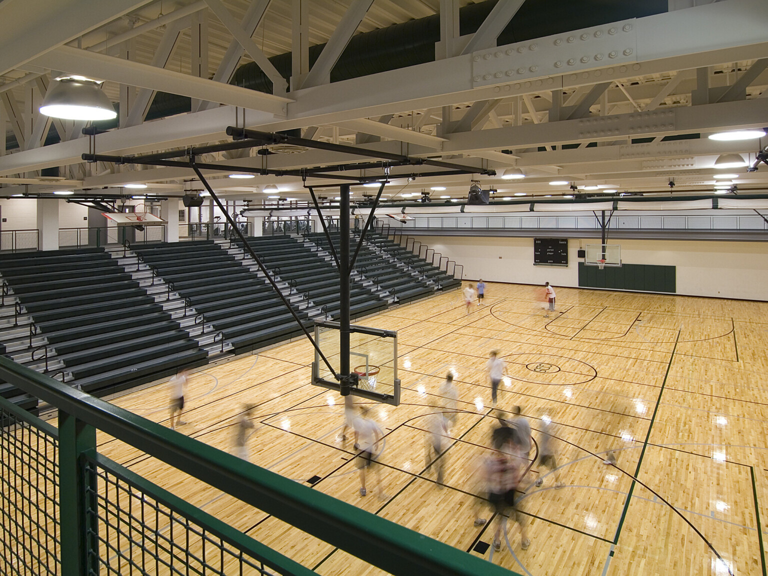 High school gymnasium with green and natural wood flooring, basketball goals suspended from the ceiling, and green bleachers to the left