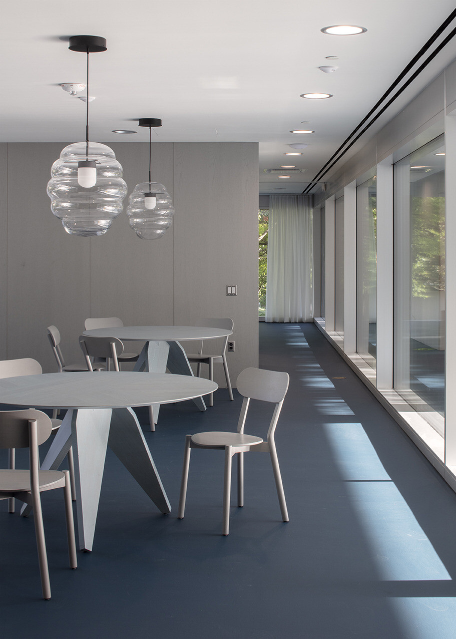 Seating area inside a building with floor to ceiling windows, blue flooring, white tables and chairs, modern glass fixtures suspended from the ceiling, grey walls