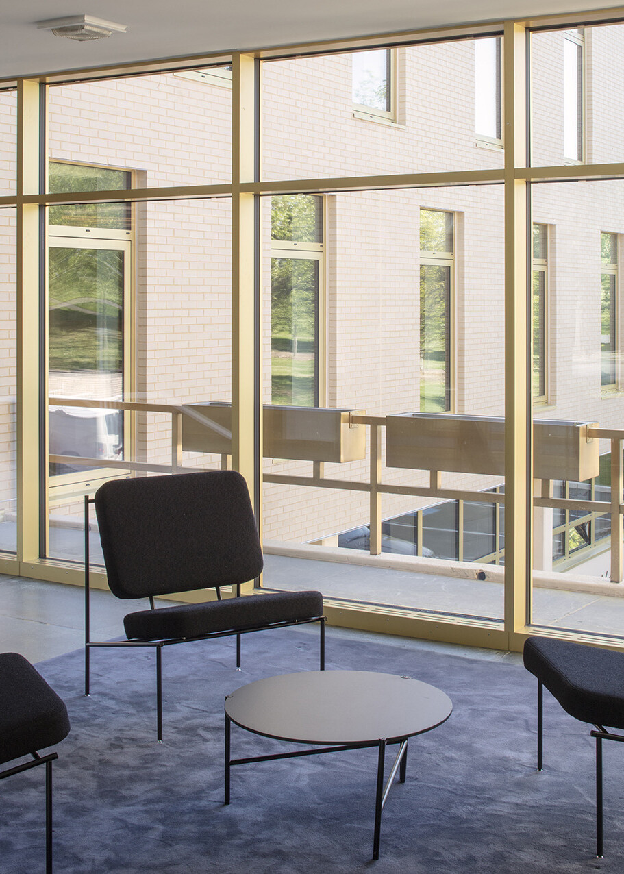 Floor to ceiling windows framed in brushed gold, modern chairs with black cushions, brick wall filled with windows outside