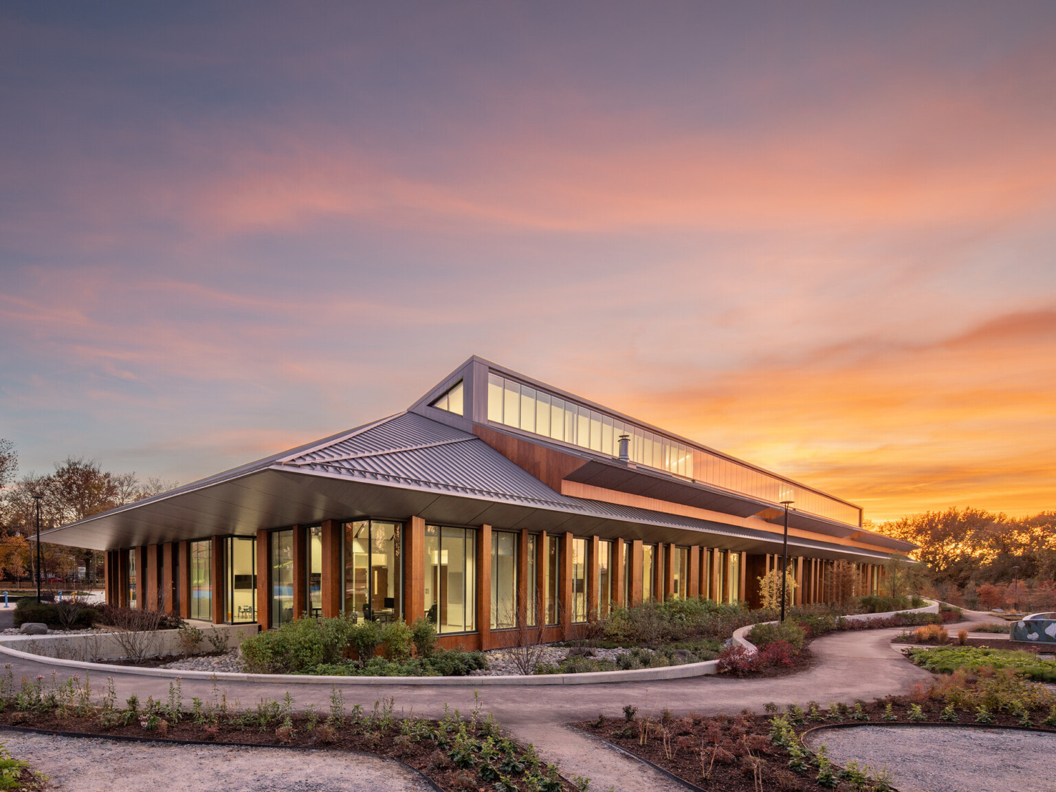 Modern, angular building with a slanted metal roof and large glass windows surrounded by lush landscaping at sunset