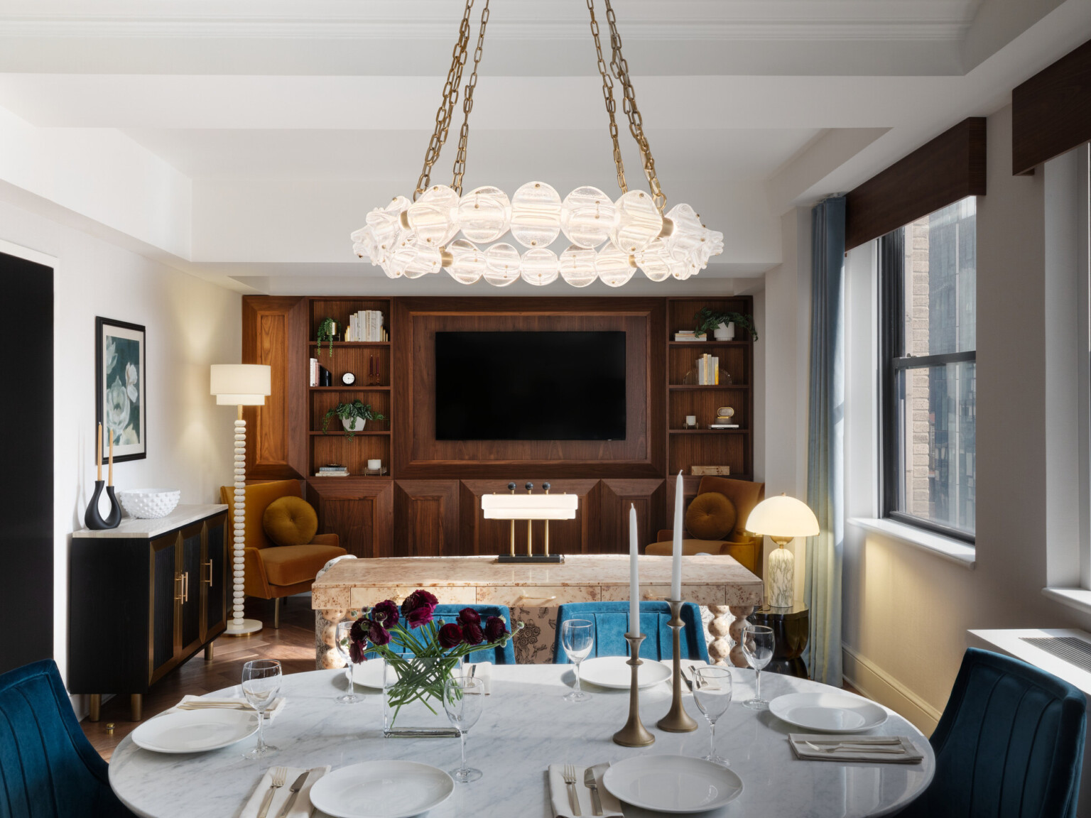 Modern living and dining spaces. Wooden wall with TV in back. White marble table surrounded by blue velvet chairs