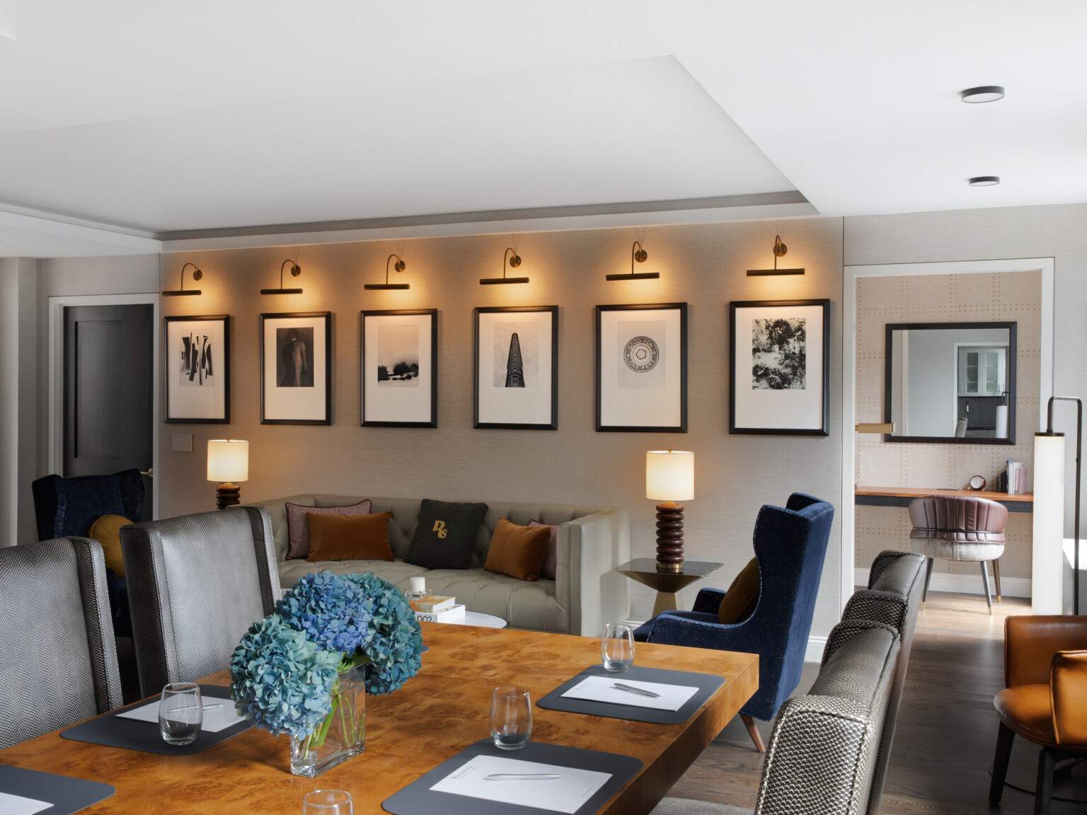 Modern living space with multiple seating areas. Rectangular table surrounded by chairs, small tufted couch and blue chairs in back with artwork displayed on wall above, doorway looking at desk space