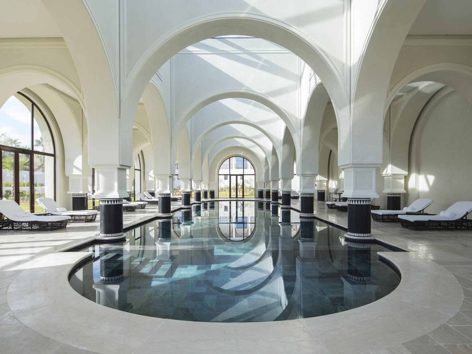 Luxurious indoor pool lined with lounge chairs. High vaulted arches throughout and columns with black bases in the pool