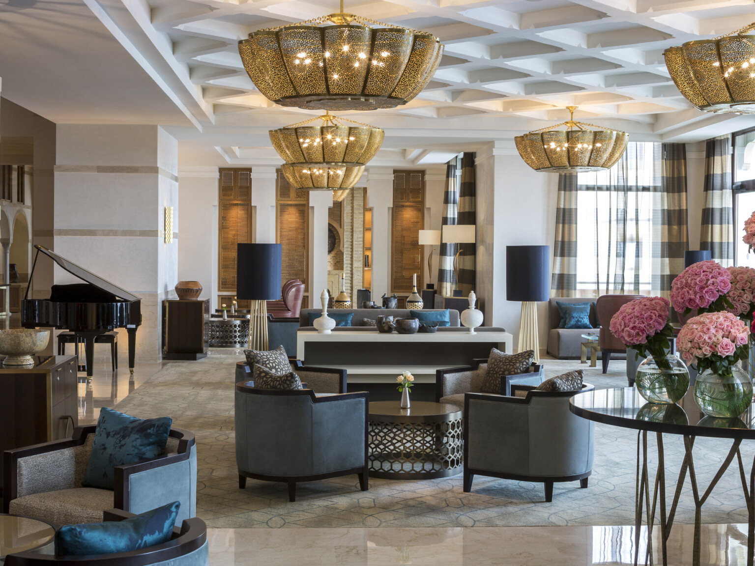 Luxurious hotel lobby with gold chandeliers, multiple seating areas, a grand piano, and vases of pink roses on a round table