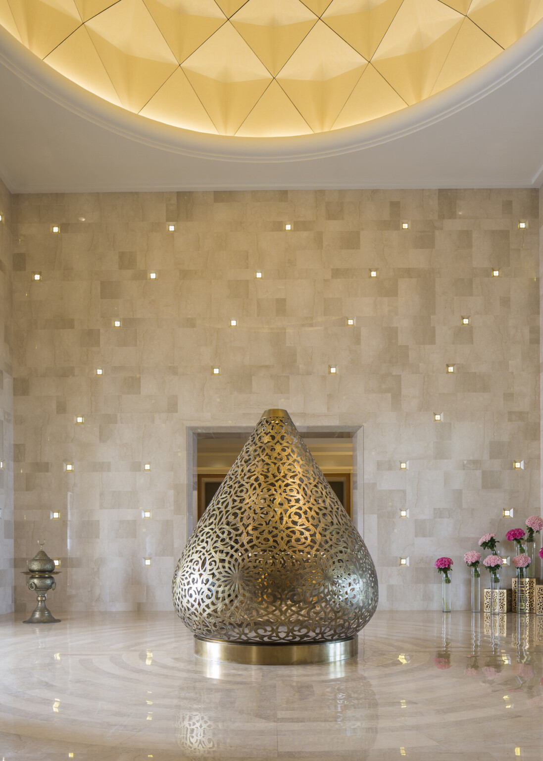 Luxurious interior space with a focus on a large, ornate metallic teardrop shaped sculpture in the center of the room. Pink flowers in glass containers in the right corner of the room