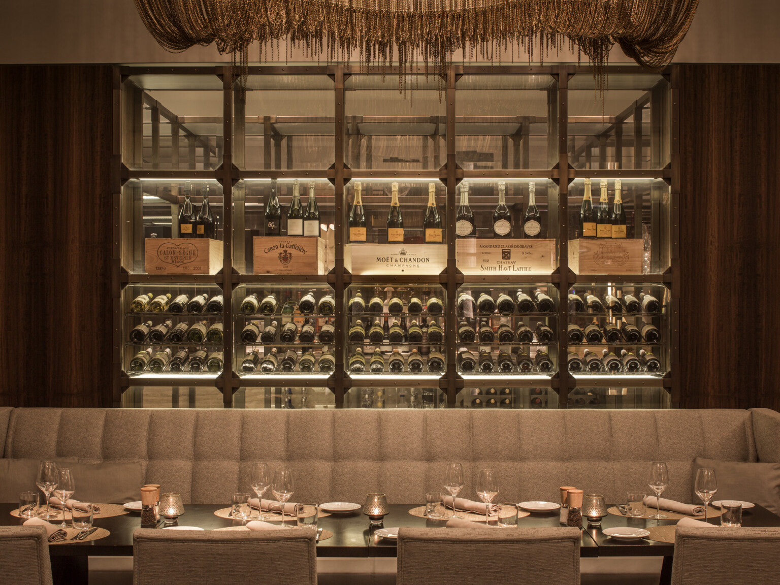 An upscale dining area with a plush bench behind an elegantly set table, and a glass wine rack in the background displaying illuminated wine bottles.