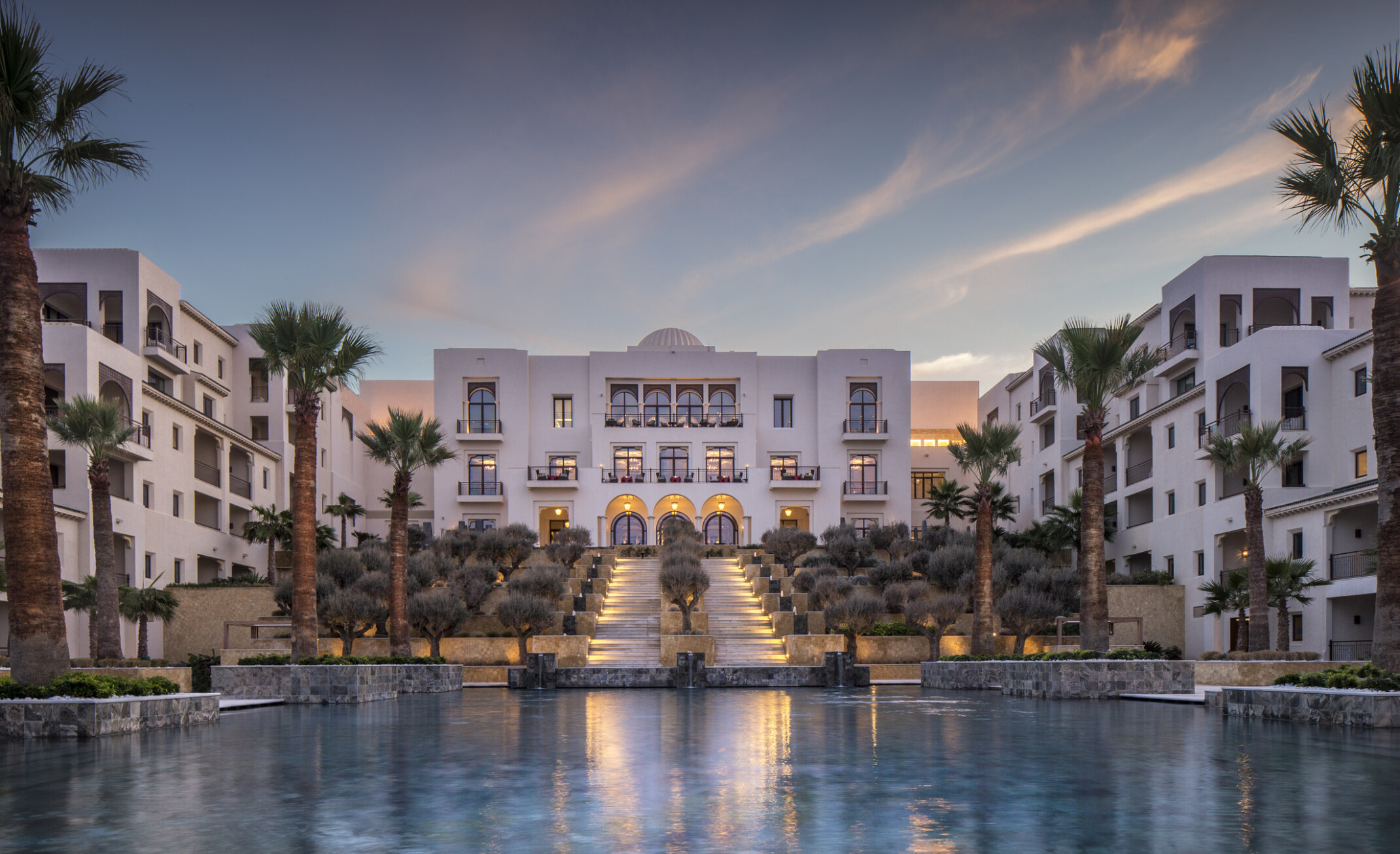 Luxurious white hotel buildings with palm trees, illuminated at dusk, and surrounding a reflective pool in front.