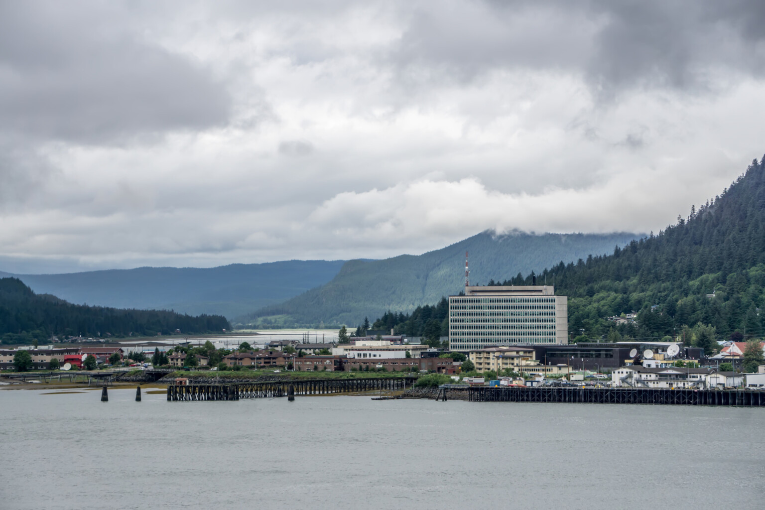 Scenic view of the Juneau Federal Building and U.S. Courthouse, with the Gastineau Channel in the foreground and fir-covered coastal hills in the background. Sky is gray with low clouds