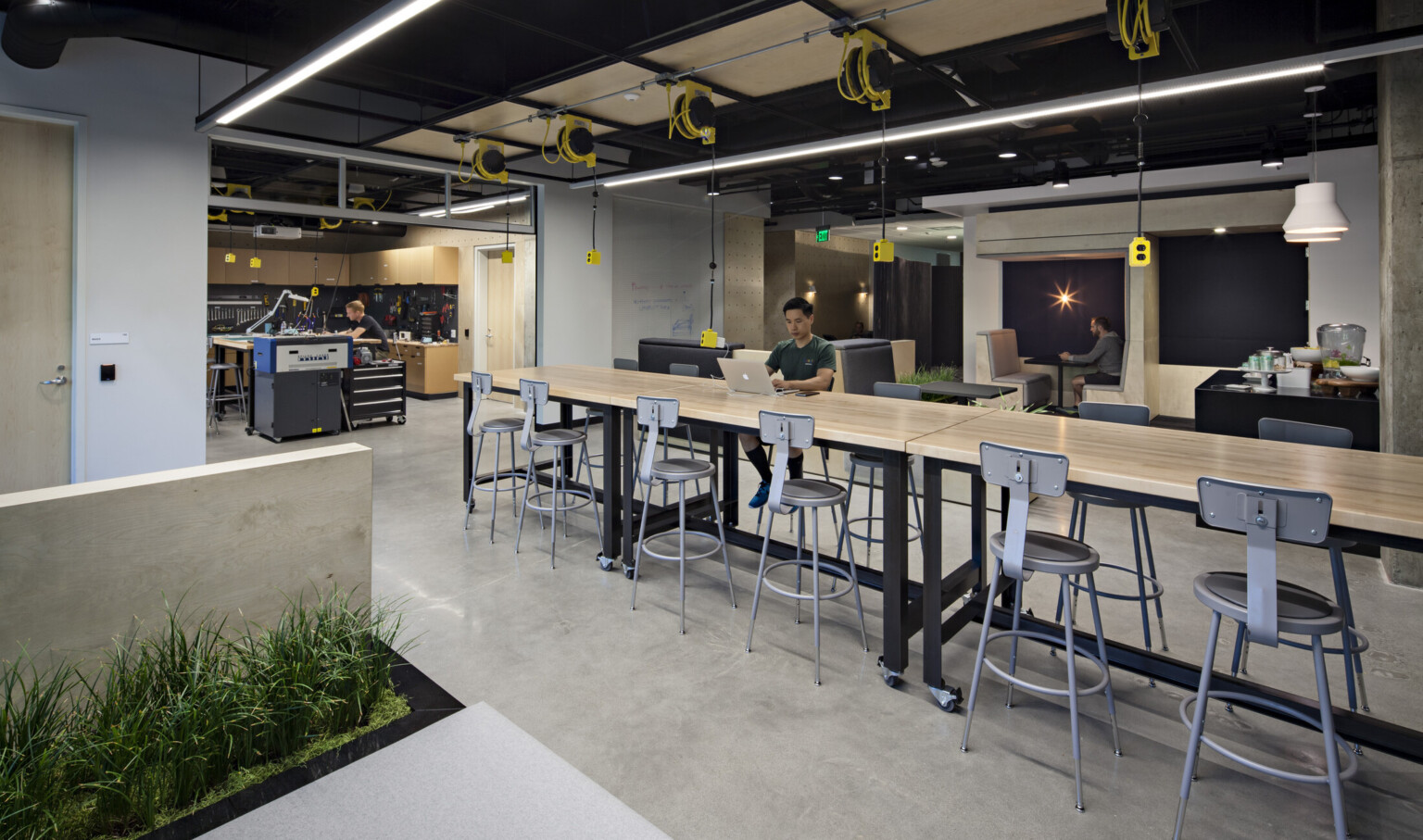 Office lounge with industrial seating surrounding a light wood table, yellow and black electrical outlets suspended from the ceiling and a workshop in the background with cabinets lining the walls and a high top table in the center of the room