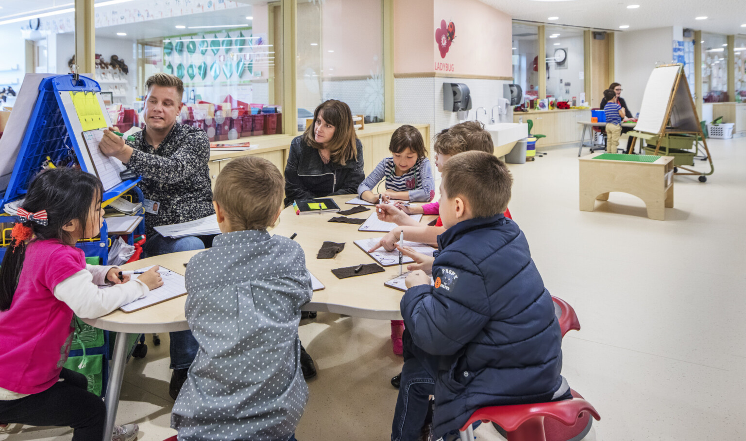 Group of children gathered around a table listening to a teacher teach off of a whiteboard